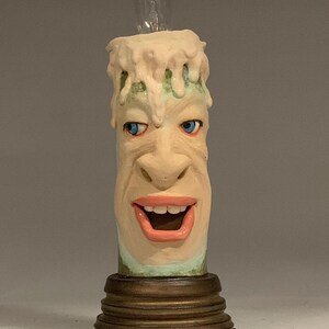 Könnte beinhalten: Eine Kerze aus Keramik mit einem auf der Vorderseite modellierten Gesicht. Die Kerze ist mit einer Glühbirne beleuchtet und hat eine grün-weiße Basis. Die Kerze steht auf einem goldenen Sockel mit mehreren Ringen.