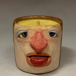 Puede incluir: Un portavelas de cerámica con diseño de rostro. La vela tiene cera amarilla y una mecha. La cara tiene ojos azules, nariz roja y lengua roja. El texto "The Art of Lucky Stradley" está en la parte inferior.