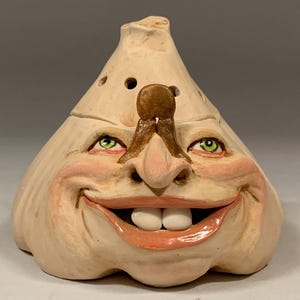 May include: A ceramic garlic-shaped sculpture with a beige finish, featuring a wide smiling face with two prominent white teeth, green eyes, and a brown nose. The top has small ventilation holes. The text The Art of Lucky Stradley is below.