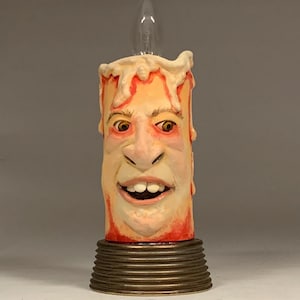 Op de afbeelding: Een unieke kaarssculptuur met een uitgesneden gezicht, met een gloeilamp aan de bovenkant. De kaars heeft een geel en rood kleurenschema, met een gedetailleerd gezicht en een bronzen voet. De tekst "The Art of Lucky Stradley" staat onderaan.