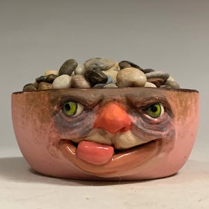 May include: A pink ceramic bowl featuring a sculpted face with green eyes, a prominent nose, and a tongue sticking out. The bowl is filled with smooth, multi-colored river stones. The text "The Art of Lucky Stradley" is printed below the bowl.
