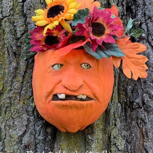 May include: A ceramic pumpkin planter with a funny face, painted orange with white teeth. The planter is decorated with a bouquet of fall flowers, including sunflowers and red and orange flowers.