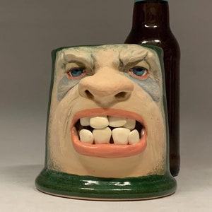 May include: A green ceramic mug with a sculpted face. The face has a wide mouth with teeth, a large nose, and blue eyes. The mug is next to a bottle of Angry Orchard hard cider.