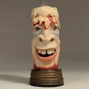 Op de afbeelding: Een unieke, handgeschilderde kaars met een gezicht in het was. De kaars heeft een grijnzende mond, blauwe en groene ogen en een gloeilamp. De basis is messingkleurig metaal. De tekst "The Art of Lucky Stradley" staat onderaan.