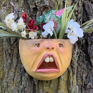 May include: A yellow ceramic planter shaped like a face with an open mouth and white teeth. The planter is filled with white and red flowers and green foliage.