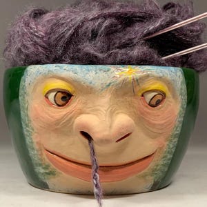Puede incluir: Un cuenco de hilo de cerámica caprichoso con un diseño de cara de mono. El cuenco es verde y azul, lleno de hilo morado y agujas de tejer. El texto "The Art of Lucky Stradley" está en la parte inferior.