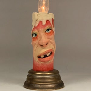 Könnte beinhalten: Eine Kerzen-Skulptur aus Keramik mit einem menschlichen Gesicht und einer leuchtenden Glühbirne als Flamme. Die Kerze ist in Rot- und Weißtönen bemalt und steht auf einem goldenen Sockel.