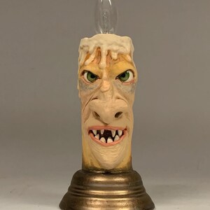 Könnte beinhalten: Eine Kerze aus Keramik mit einem grotesken Gesicht und tropfendem Wachs, beleuchtet von einer Glühbirne. Die Kerze steht auf einem goldenen Sockel.