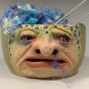 Puede incluir: Un cuenco de hilo de cerámica con diseño de rostro, lleno de hilo azul y blanco y dos agujas de tejer plateadas. El cuenco tiene un aspecto envejecido y desgastado con un rostro detallado y el texto "The Art of Lucky Stradley".