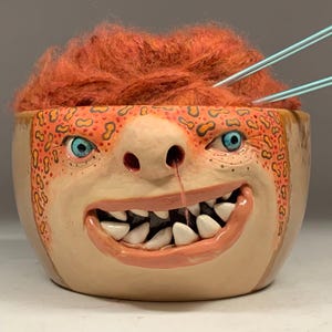 Könnte beinhalten: Eine skurrile Keramikschale mit einem Monstergesicht-Design. Die Schale hat blaue Augen, ein breites Grinsen mit weißen Zähnen und orangefarbenes Haar. Zwei Stricknadeln sind in das orangefarbene Haar gesteckt. Der Text "The Art of Lucky Stradley" befindet sich unten.