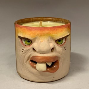 Puede incluir: Un portavelas de cerámica con forma de cara con una amplia sonrisa de dientes. El portavelas está pintado en tonos de marrón, naranja y amarillo, con ojos verdes y un diente blanco. El portavelas está lleno de una vela blanca.