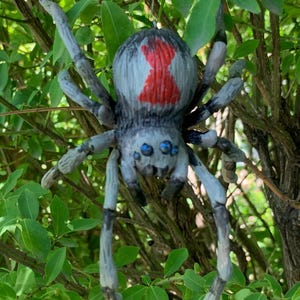 May include: A gray and black spider ornament with a red hourglass marking on its back, hanging among green leaves and branches. The spider has blue eyes and is suspended by a string. The text "The Art of Lucky Stradley" is at the bottom.