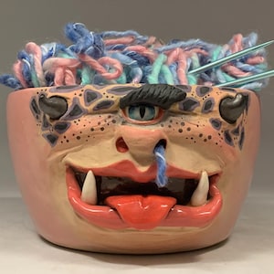 Puede incluir: Un cuenco de cerámica rosa con diseño de cara de monstruo, lleno de hilo colorido y agujas de tejer. El cuenco presenta una cara detallada con un solo ojo, dientes y una lengua. El texto "The Art of Lucky Stradley" está en la parte inferior.