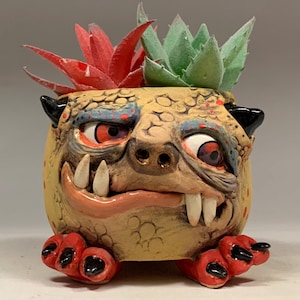May include: A quirky ceramic monster face planter, hand-painted with tan, red, blue, and black details. It has large teeth and red eyes, and is planted with two artificial succulents, one red and one green. The text "The Art of Lucky Stradley" is at the bottom.