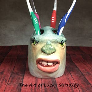 May include: A ceramic toothbrush holder with a face design, holding four toothbrushes with green, red, and blue bristles. The holder has a light green and beige color scheme, with the text "The Art of Lucky Stradley" at the bottom.