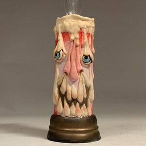 May include: A ceramic candle with a dripping wax design and a face sculpted on the front. The candle has a light bulb inside and is on a gold base.
