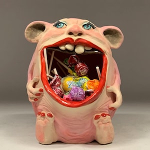 May include: A pink, monster-like ceramic sculpture with a wide open mouth filled with colorful candies. The sculpture has large ears, blue eyes, and red lips. The text "The Art of Lucky Stradley" is at the bottom.