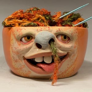 Könnte beinhalten: Eine Keramikschale mit Gesichtsdesign, gefüllt mit orangefarbenem und grünem Garn und zwei Stricknadeln. Die Schale hat eine hellorangefarbene Außenseite und ein Gesicht mit Nase, Augen und herausgestreckter Zunge. Der Text "The Art of Lucky Stradley" steht unten.