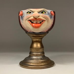 May include: A ceramic goblet with a hand-painted face. The goblet is painted in shades of red, white, and blue, with green eyes and a red mouth. The goblet is sitting on a gold-colored base.
