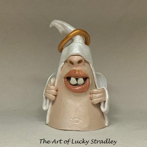 Può includere: Una statuetta di angelo in ceramica bianca con un'aureola dorata, che indossa una veste bianca e un ampio sorriso con denti grandi. L'angelo tiene la veste davanti al suo viso.