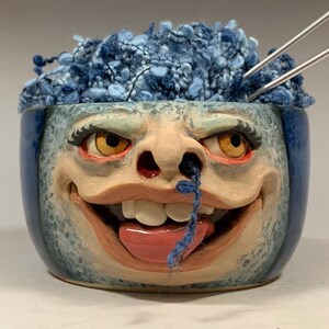 Puede incluir: Un cuenco de cerámica con un diseño de rostro caprichoso, lleno de lana azul y agujas de tejer. El cuenco presenta una cara sonriente con la lengua fuera y un mechón de hilo azul. El texto "The Art of Lucky Stradley" está en la parte inferior.
