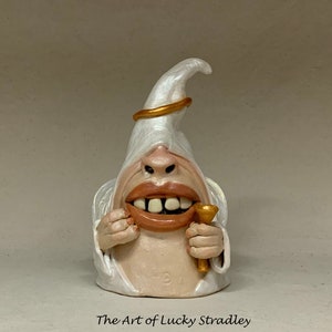 Può includere: Una statuetta di angelo in ceramica bianca con un'aureola dorata, che tiene una tromba dorata. L'angelo ha un ampio sorriso con i denti e indossa una veste bianca.