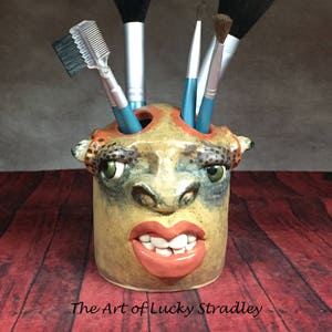 May include: A ceramic brush holder shaped like a cartoon face with a toothy grin and bright red lips. The holder has five slots for brushes and is decorated with brown, orange, and green paint.