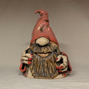 May include: A ceramic gnome figurine with a pink hat, a long brown beard, and red painted fingernails. The gnome is smiling and has a white nose.