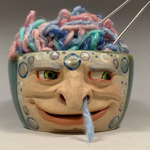 Puede incluir: Un cuenco de hilo de cerámica caprichoso con un diseño de cara, que contiene hilo colorido y agujas de tejer. El cuenco está pintado con una base beige claro, detalles turquesa y ojos verdes. El texto "The Art of Lucky Stradley" está en la parte inferior.