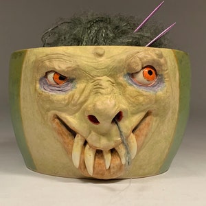 Könnte beinhalten: Eine Keramik-Garn-Schale in Form eines Monstergesichts mit grüner und gelber Farbe. Das Monster hat orange Augen, eine große Nase und scharfe Zähne. Die Schale ist mit grauem Garn und zwei lila Stricknadeln gefüllt.