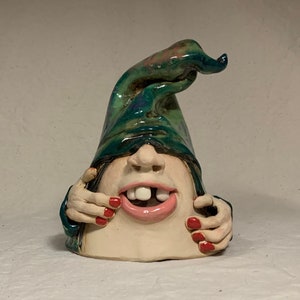 Può includere: Una statuetta in ceramica di una persona con un cappello verde tirato sugli occhi. La persona ha un ampio sorriso con un dente mancante e smalto per unghie rosso sulle dita.
