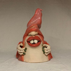 Può includere: Una scultura in ceramica di una persona con una grande bocca aperta e un cappello rosa. La persona indossa una veste rossa e ha le unghie verdi.