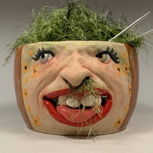 Puede incluir: Una maceta de cerámica con diseño de rostro. La maceta tiene un borde rojo, una cara con labios rojos y detalles de flores amarillas. Material vegetal verde y agujas de tejer en el interior. El texto "The Art of Lucky Stradley" está en la parte inferior.