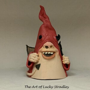 May include: A small ceramic sculpture of a devil with a red pointed hat, a mischievous grin, and a tail. The sculpture is painted in shades of red, black, and white.