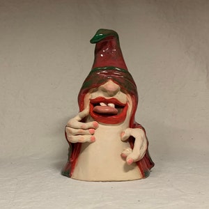 May include: A ceramic figurine of a whimsical character with a red and green hat, a wide smile, and a large tongue sticking out. The character is wearing a red robe and has pink fingernails.