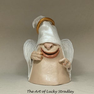 May include: A white ceramic angel figurine with a gold halo, smiling with a wide open mouth. The angel is wearing a white robe and has wings.