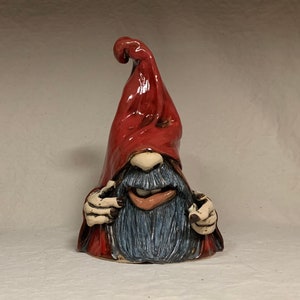 May include: A ceramic gnome figurine with a red hat and a blue beard. The gnome is smiling and has a mischievous expression. The gnome is handmade and has a unique, whimsical style.