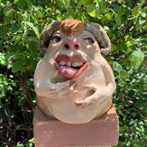 Puede incluir: Una escultura de cerámica caprichosa de una criatura con cuernos, ojos grandes y una lengua que sobresale. La escultura está pintada en tonos beige, marrón y rosa, y está montada sobre una base cuadrada. Fondo verde.