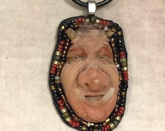 Pode incluir: Um colar com pingente com um rosto esculpido com uma expressão sorridente. O pingente é emoldurado com contas pretas, vermelhas e douradas e pende de um cordão preto. O texto "The Art of Lucky Stradley" está na parte inferior.