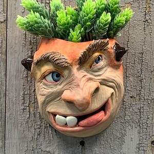 May include: A ceramic wall planter in the shape of a grinning face with blue eyes and a protruding tongue, filled with faux green plants. The planter has a textured, aged finish. The text "The Art of Lucky Stradley" is visible at the bottom.