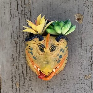 May include: A ceramic planter shaped like a cartoon monster's face, painted in yellow, orange, and black. The planter is filled with green and yellow succulents.