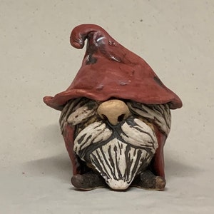 May include: A small ceramic gnome figurine with a red hat and a white beard. The gnome is sitting with its hands crossed in front of it.