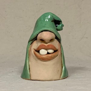 Può includere: Una figurina in ceramica fantasiosa di un viso con un cappello verde. Il viso ha un ampio sorriso con due grandi denti bianchi. La figurina è realizzata in stile folk.