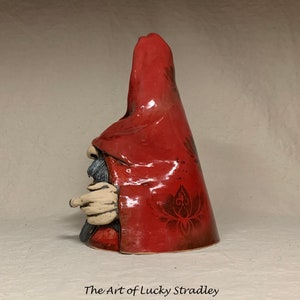 May include: A ceramic gnome figurine with a red hooded cloak and a detailed floral pattern. The gnome has a long white beard and is holding a finger to its lips.