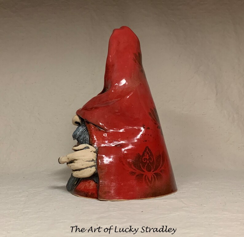 May include: A ceramic gnome figurine with a red hooded cloak and a detailed floral pattern. The gnome has a long white beard and is holding a finger to its lips.