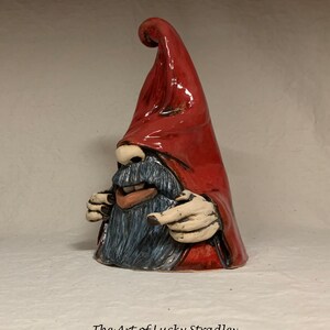 May include: A ceramic gnome figurine with a red pointed hat, a blue beard, and a mischievous expression. The gnome is pointing with both hands.