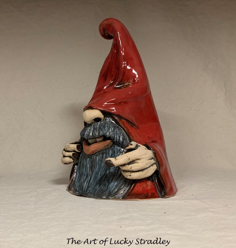 May include: A ceramic gnome figurine with a red pointed hat, a blue beard, and a mischievous expression. The gnome is pointing with both hands.