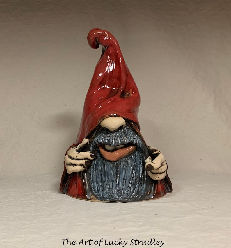 May include: A ceramic gnome figurine with a red hat and a blue beard. The gnome is smiling and has a mischievous expression. The gnome is handmade and has a unique, whimsical style.