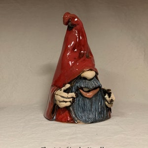 May include: A ceramic gnome figurine with a red pointed hat, a blue beard, and a whimsical expression. The gnome is holding a small object in its hand.
