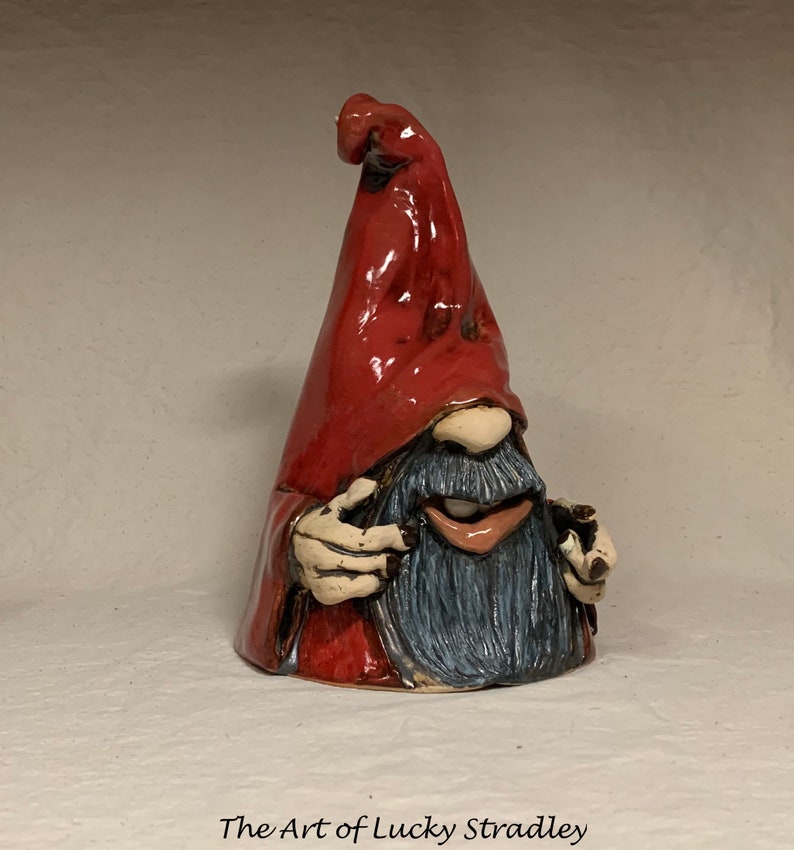 May include: A ceramic gnome figurine with a red pointed hat, a blue beard, and a whimsical expression. The gnome is holding a small object in its hand.